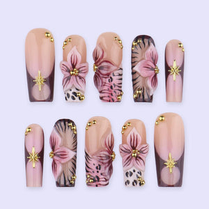 A set of ten artificial nails in various designs. The nails feature a nude base with floral and animal print accents, gold embellishments, and starburst or floral designs. The nails are in a coffin shape.