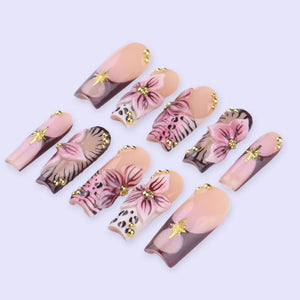 A collection of ten artificial nails with a variety of designs. The nails feature a combination of light pink, brown, and gold accents. Some nails have floral designs, while others have gold star embellishments.