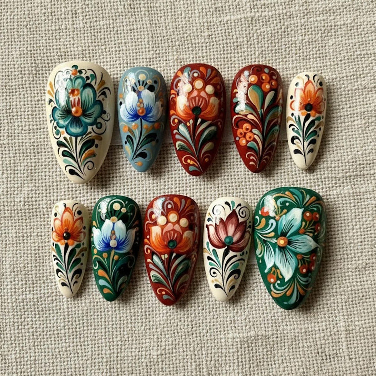 A collection of ten painted artificial nails, each with a unique floral design. The nails feature a variety of colors, including white, red, green, blue, and orange, with intricate patterns and details. The nails are arranged on a neutral fabric surf