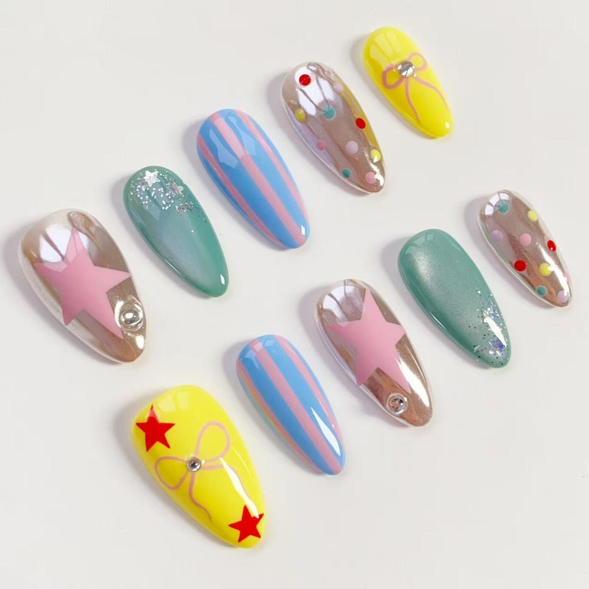 A collection of artificial nails in various designs. The nails feature a range of colors, including yellow, blue, pink, and green. Some nails have star, stripe, and dot patterns, while others have glitter or bow accents. Each nail has a glossy finish