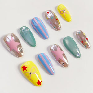 A collection of artificial nails in various designs. The nails feature a range of colors, including yellow, blue, pink, and green. Some nails have star, stripe, and dot patterns, while others have glitter or bow accents. Each nail has a glossy finish