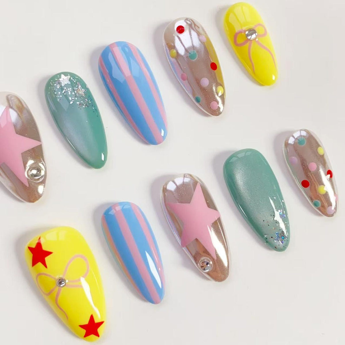 A collection of artificial nails with various designs. Some nails are yellow with red stars and a bow, others are blue with pink stripes, and some have pink stars or colorful polka dots. The nails are oval-shaped and have a glossy finish.