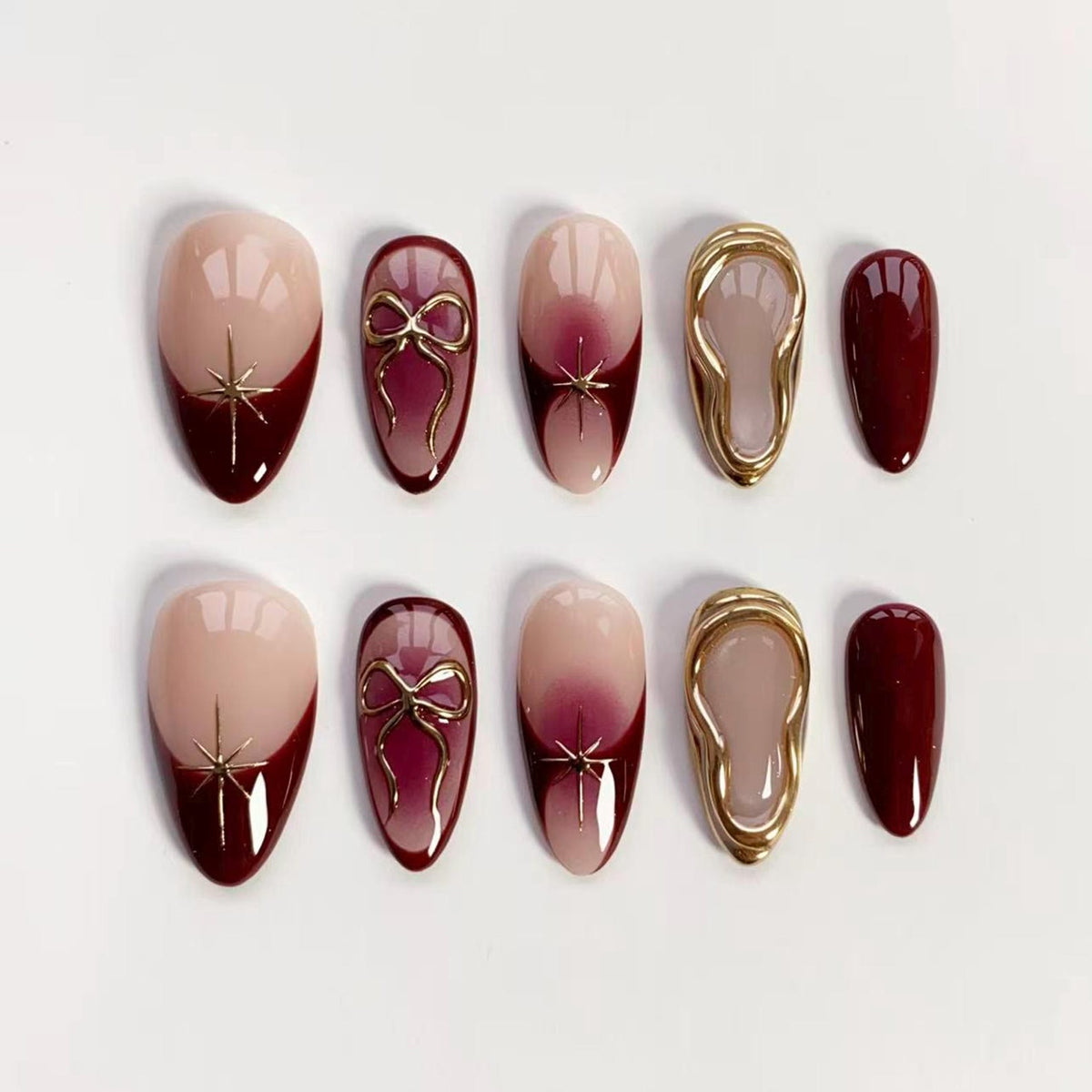 A set of ten artificial nails in various designs. Some nails are nude with a dark red tip and gold starburst, others feature a gold bow, and some have a gold frame. The nails are oval-shaped and have a glossy finish.