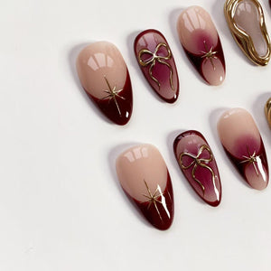 A collection of almond-shaped artificial nails. The nails feature a gradient design, transitioning from a nude pink to a deep burgundy. Some nails have gold starburst accents, while others showcase gold bow embellishments.