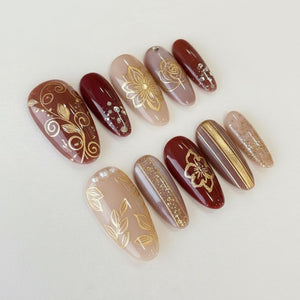 A collection of artificial nails in various shades of brown, burgundy, and beige. Each nail features intricate gold designs, including floral patterns, swirls, and glitter accents. The nails are arranged on a white surface.