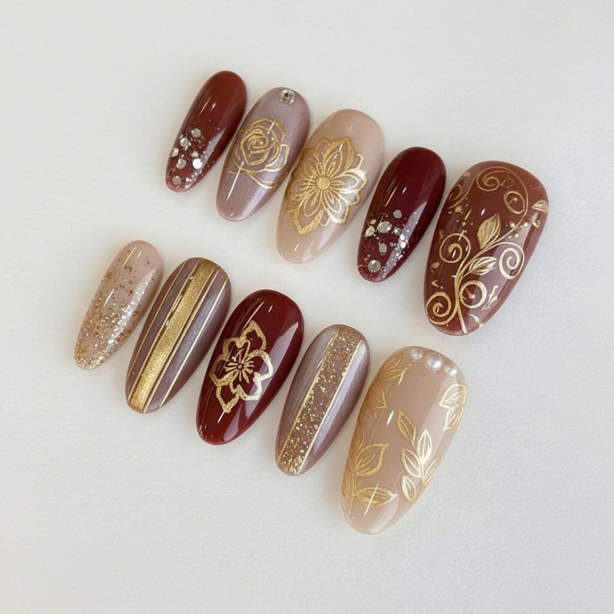 A collection of ten artificial nails with various designs. The nails feature a range of colors, including burgundy, nude, and gold. Some nails have gold floral patterns, glitter accents, or metallic stripes. The nails are oval-shaped.
