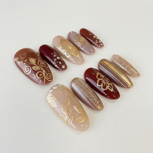 A collection of elegant, almond-shaped artificial nails. The nails feature various designs, including gold floral patterns, glitter accents, and stripes, set against a palette of burgundy, beige, and brown hues. Some nails have small, sparkling embel