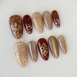 A collection of ten artificial nails in various shades of brown, beige, and burgundy. Each nail features intricate gold designs, including floral patterns, leaves, and stripes. Some nails are adorned with small, sparkling embellishments.