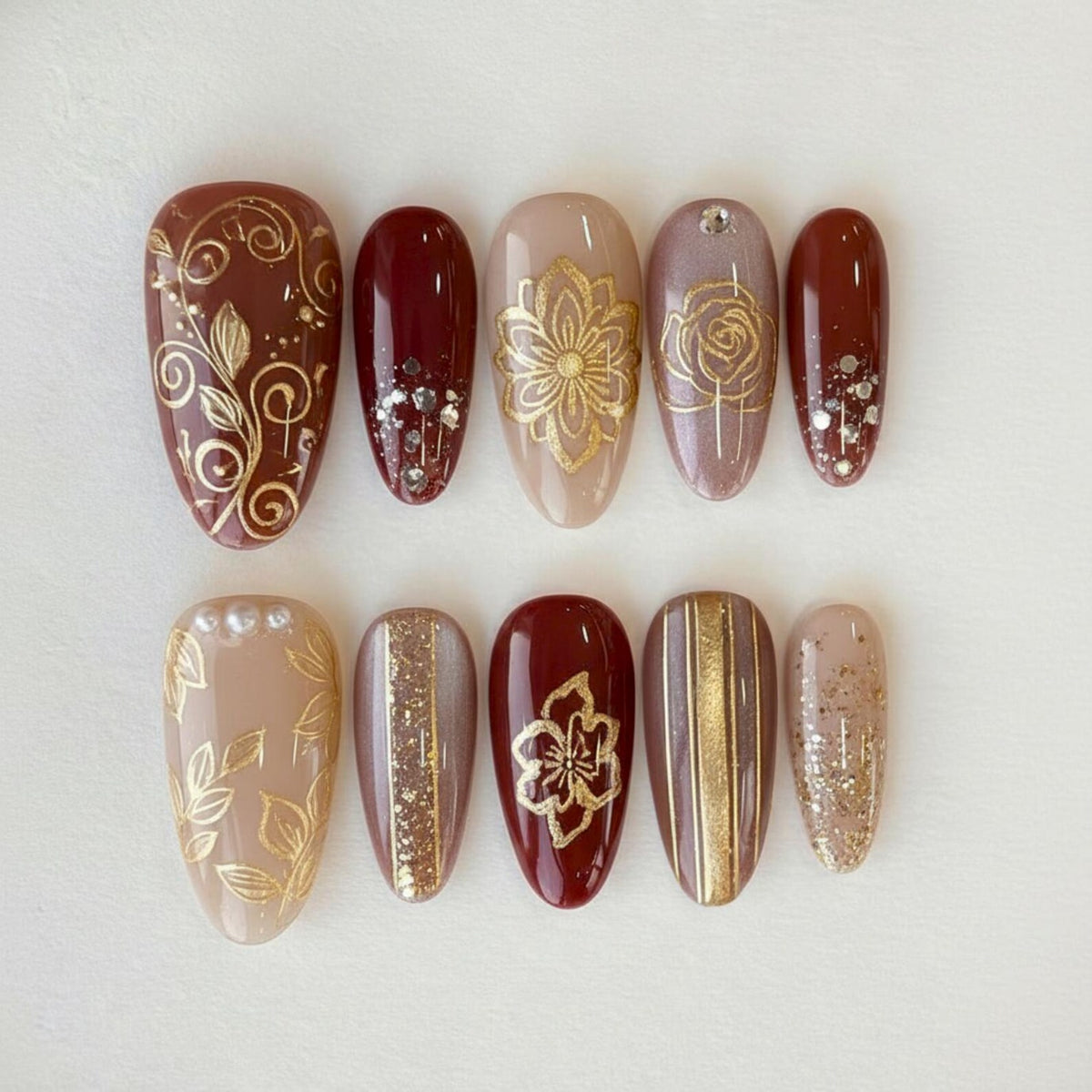 A set of ten artificial nails in various shades of brown, beige, and burgundy. Each nail features intricate gold designs, including floral patterns, swirls, and glitter accents. The nails are oval-shaped and arranged on a white surface.