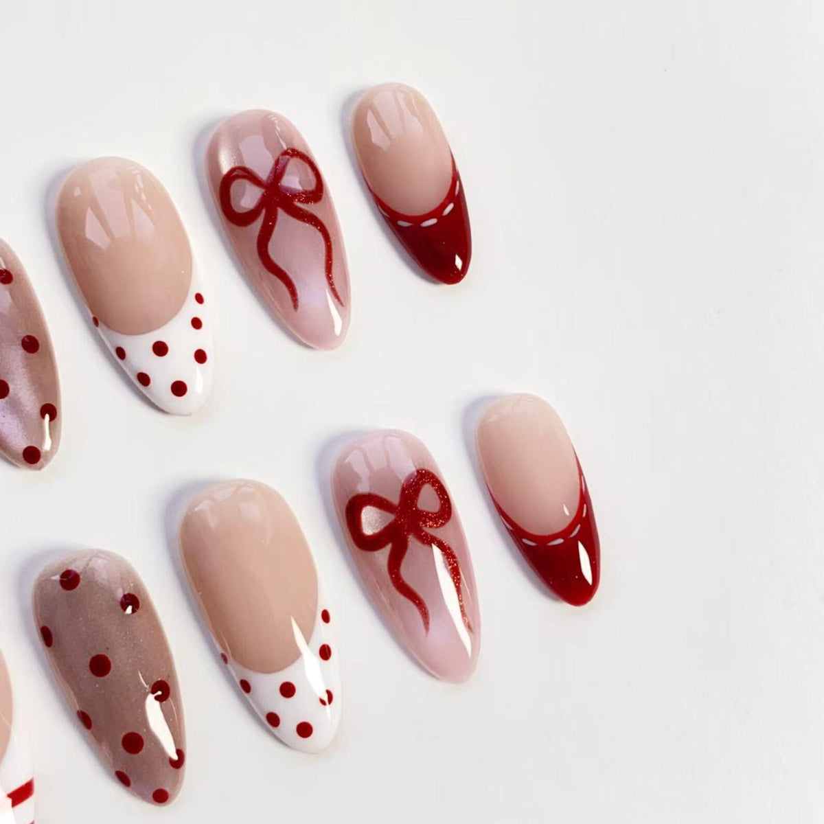 A collection of oval-shaped false nails. Some nails feature a nude base with red polka dots or a red French tip. Others have a nude base with a red bow design. The nails are arranged on a white surface.