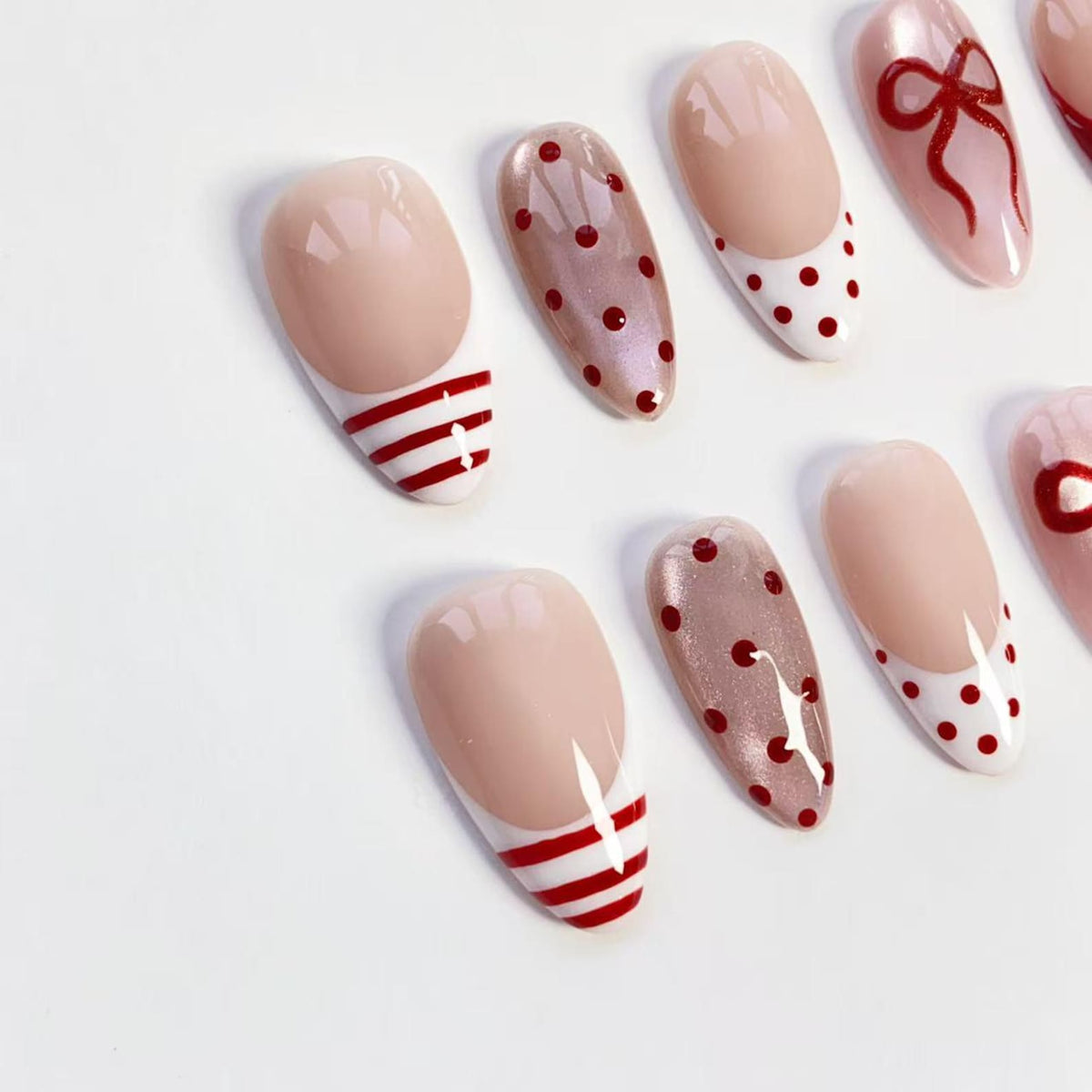 A collection of artificial nails in various designs. Some nails feature a nude base with red and white stripes, while others have a nude base with red polka dots or a white base with red polka dots. One nail has a red bow design.