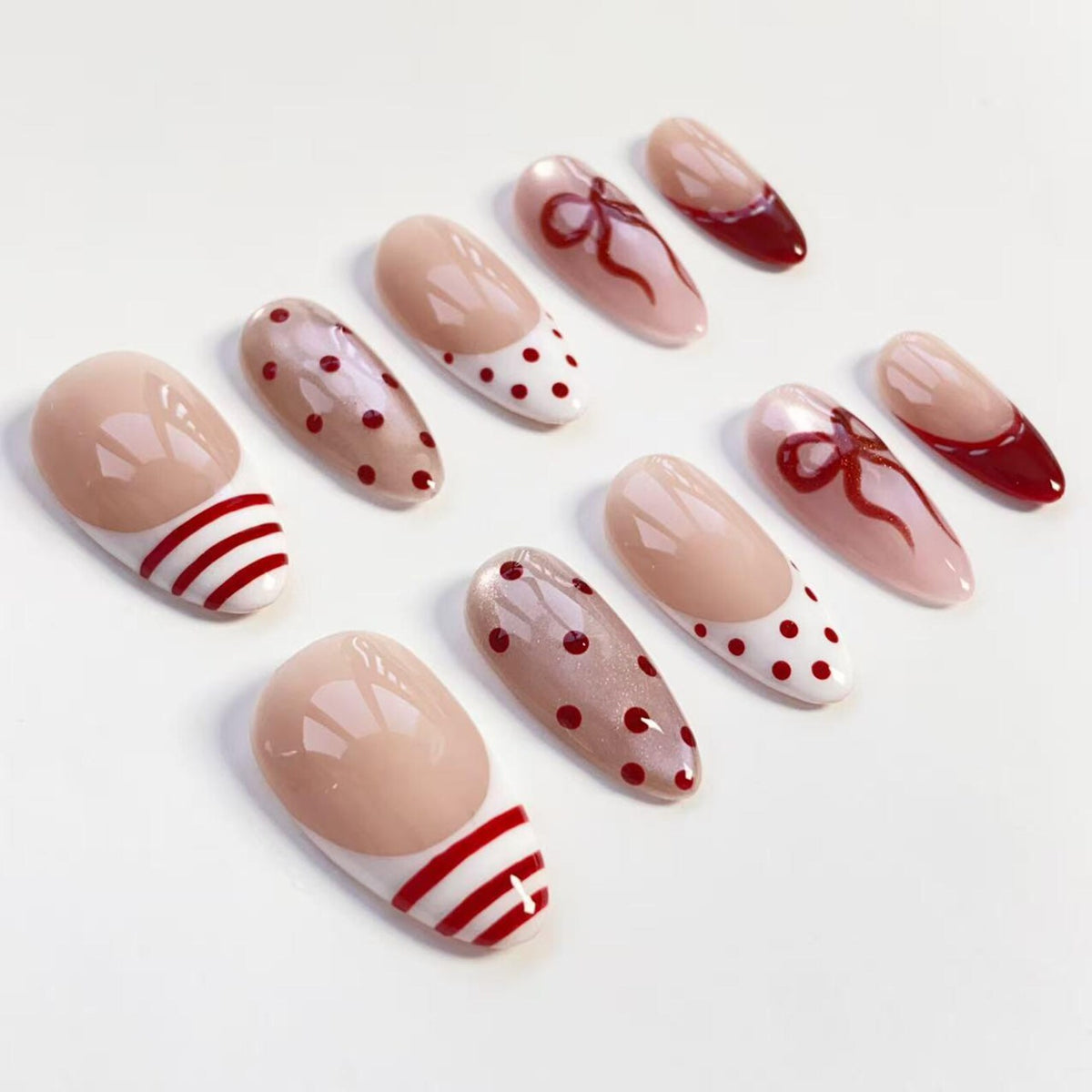A collection of artificial nails in various designs. Some nails feature a nude base with red polka dots, stripes, or bows. Others have a solid red tip. The nails are oval-shaped and arranged on a white surface.