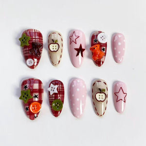 A set of ten artificial nails in various designs. Some nails feature a plaid pattern, while others are solid pink with white polka dots. Decorations include stars, buttons, and small apple and teddy bear charms.