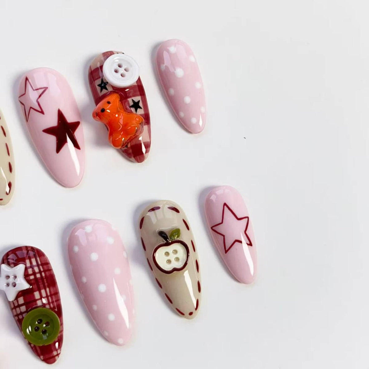 A collection of almond-shaped false nails with various designs. Some nails are pink with white polka dots, others feature star outlines, a teddy bear, or an apple with button details. The nails have a glossy finish.