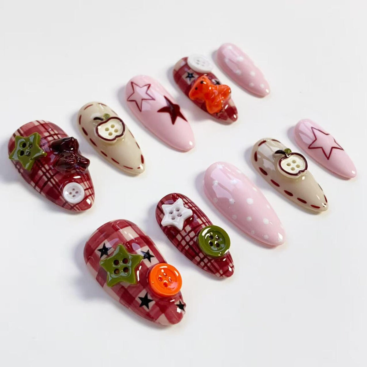 A collection of decorative acrylic nails in various designs. Some nails feature plaid patterns, star and apple accents, and colorful buttons. Others are solid pink with white polka dots or star outlines. The nails are oval-shaped.