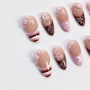 A collection of artificial nails with various designs. Some nails feature a nude base with white and brown floral patterns, while others have a nude base with white and brown stripes. Some nails have pearl accents.