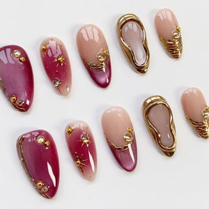 A collection of artificial nails in various designs. Some nails are a deep burgundy with gold accents and rhinestones. Others feature a gradient from pink to burgundy with starburst and gold details. Several nails have a gold frame.