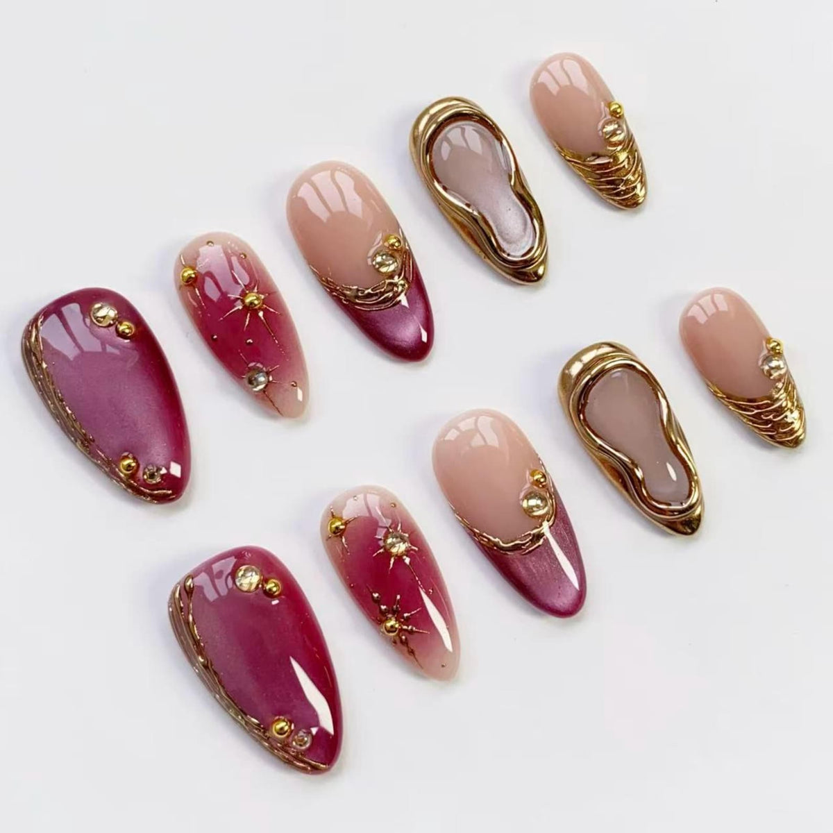 An assortment of artificial nails in various designs. Some nails are a gradient of pink and burgundy with gold accents and starburst details. Other nails feature a gold frame with a nude interior. The nails are arranged on a white surface.