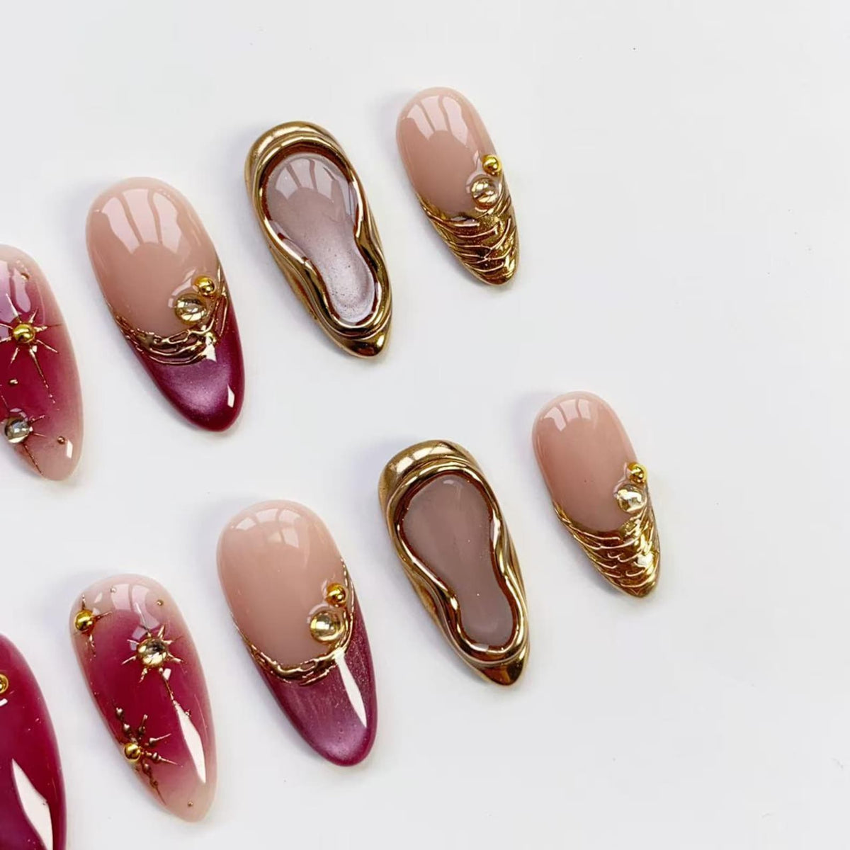 A collection of artificial nails in various designs. Some nails are a gradient of pink and burgundy with gold starburst accents. Others feature gold embellishments and a gold border. Two nails have a gold shoe design.
