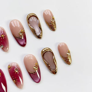 A collection of artificial nails in various designs. Some nails are a gradient of pink and burgundy with gold starburst accents. Others feature gold embellishments and a gold border. Two nails have a gold shoe design.