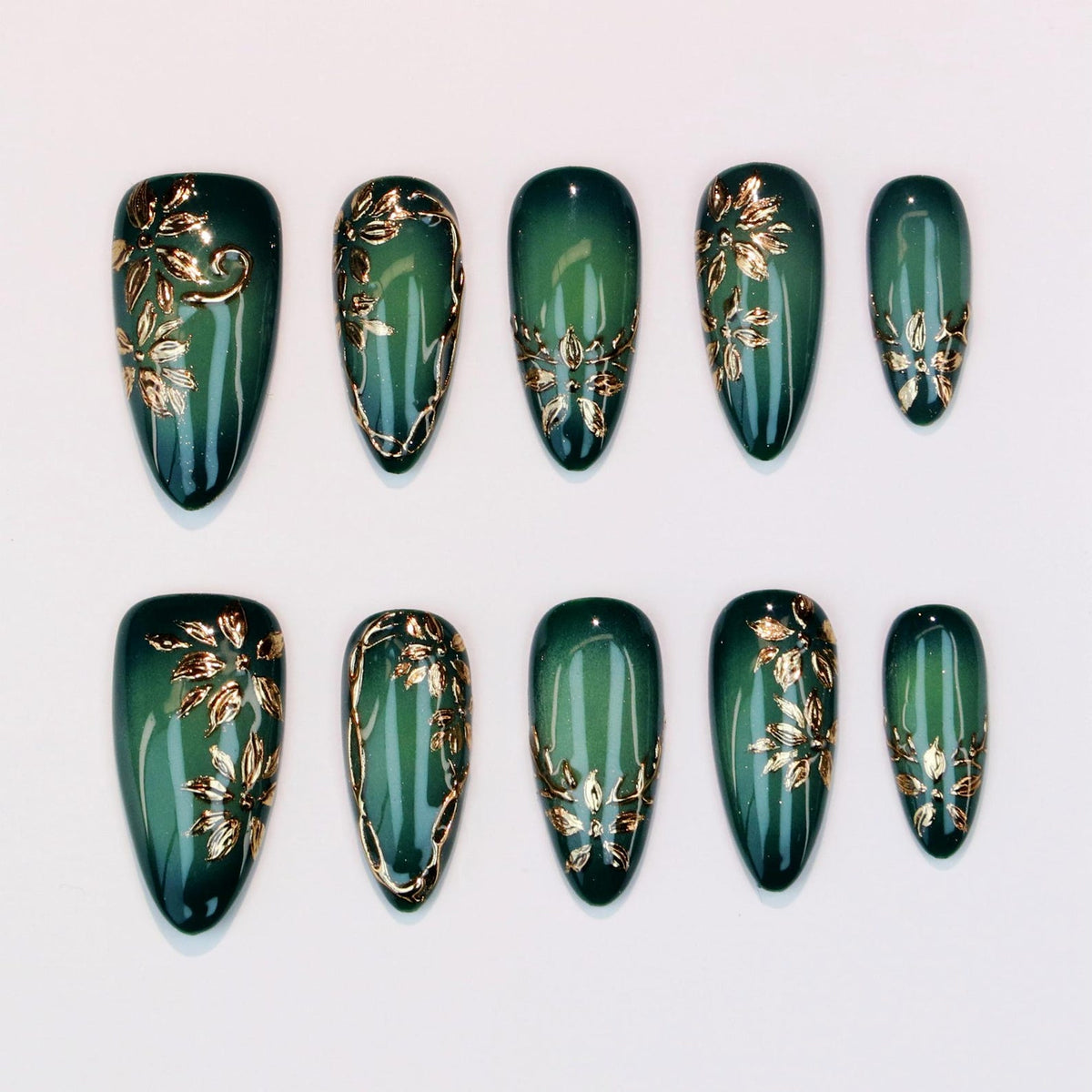Set of ten emerald green press-on nails with gold floral designs. Each nail features a glossy finish and a different gold leaf pattern. The nails are oval-shaped and arranged on a white surface.