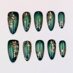 Set of ten emerald green press-on nails with gold floral designs. Each nail features a glossy finish and a different gold leaf pattern. The nails are oval-shaped and arranged on a white surface.