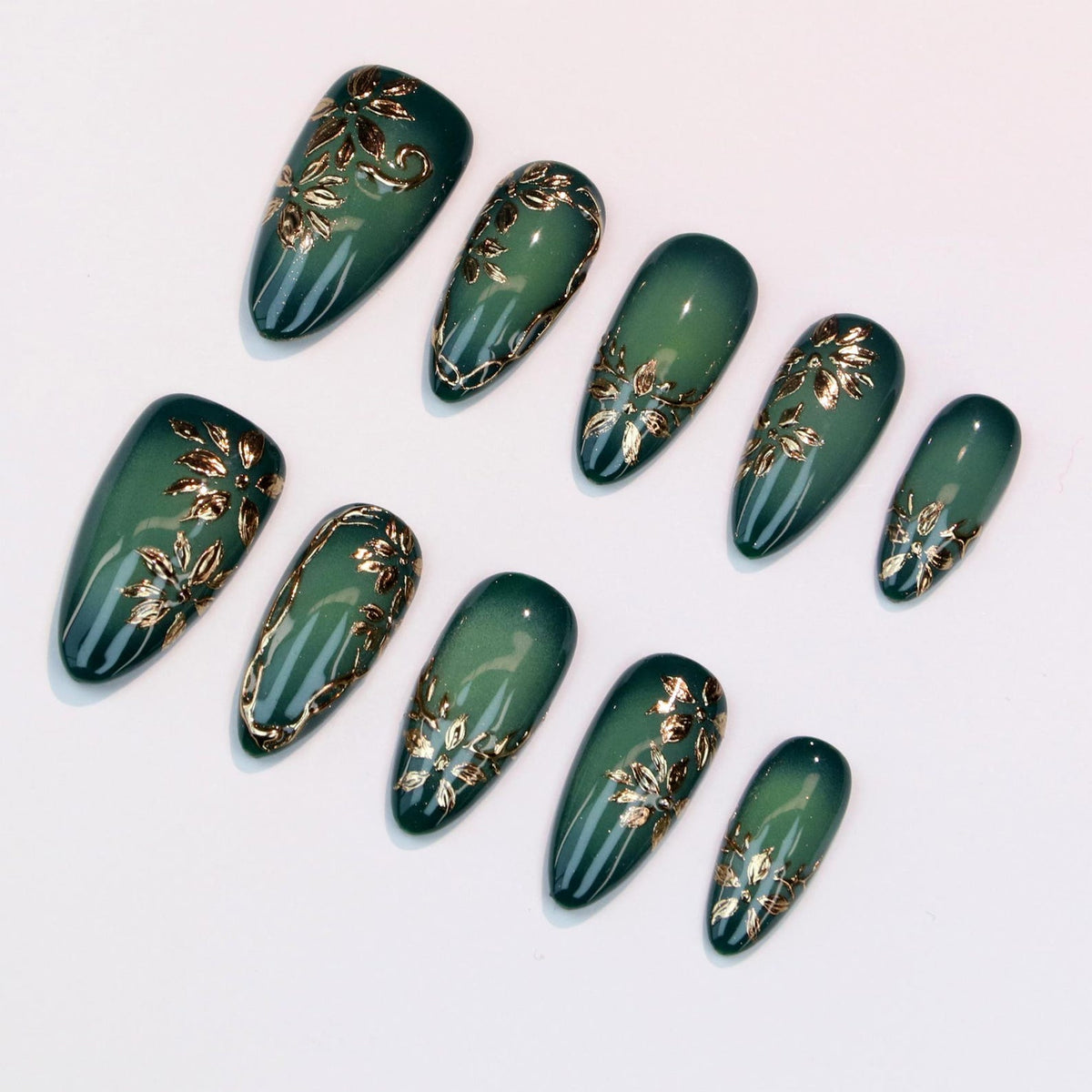 Set of ten artificial nails in various sizes. Each nail is a deep green with a gold floral design. The nails have a glossy finish and are arranged on a white surface.