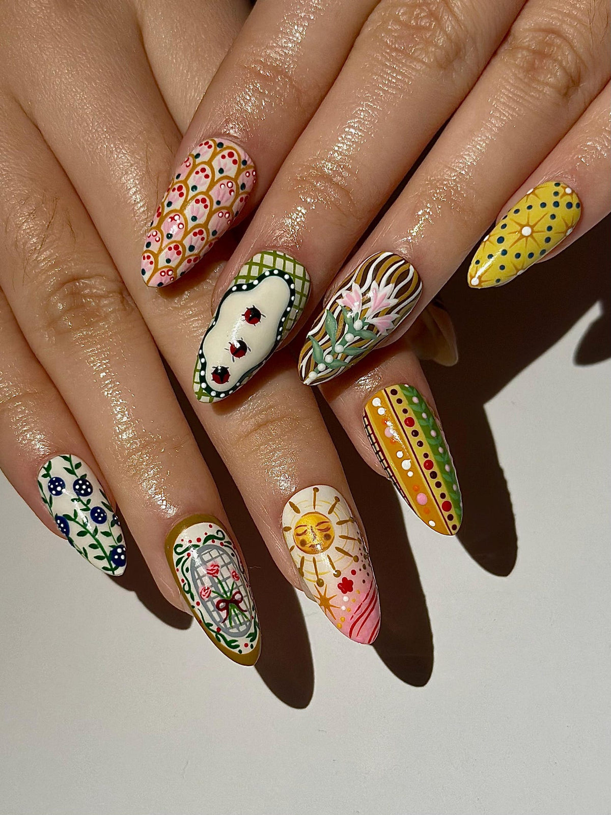 A set of long, almond-shaped nails, each painted with unique designs. The designs include ladybugs, floral patterns, suns, and geometric shapes in a variety of colors, including pink, yellow, green, and white. The nails are well-manicured and showcas