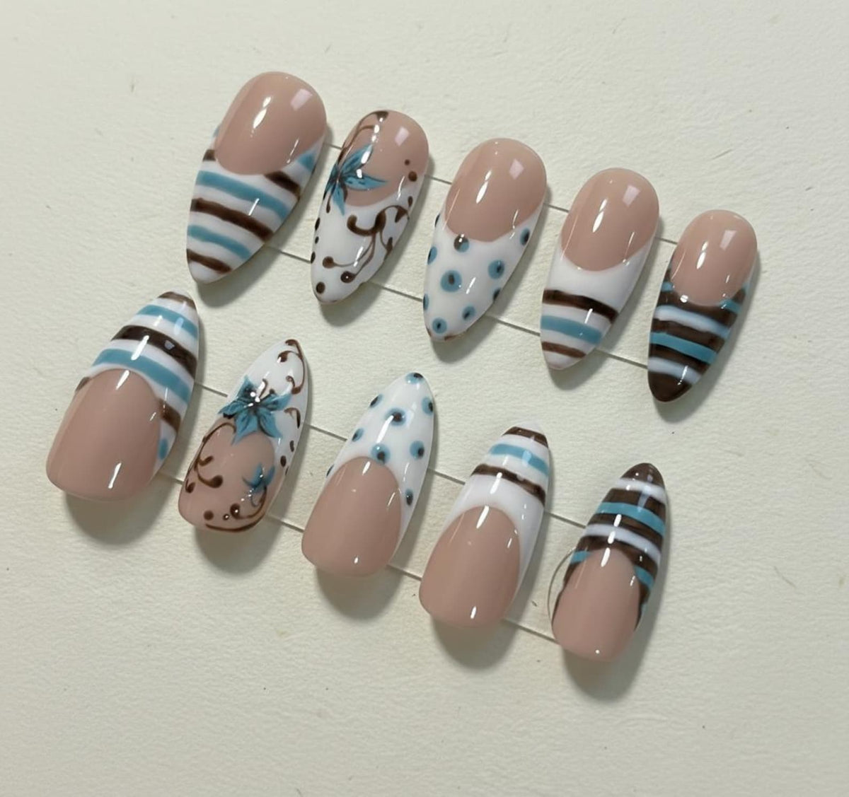 A set of ten artificial nails in various designs. Some nails feature a nude base with blue and brown stripes, while others have floral or polka dot patterns. The nails are oval-shaped and have a glossy finish.
