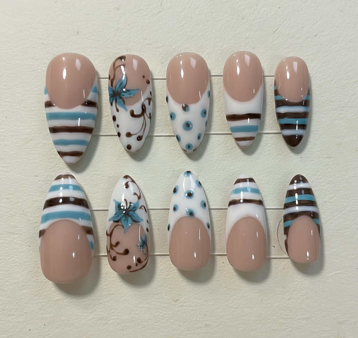 A set of ten artificial nails in various designs. Some nails feature a nude base with white, brown, and blue stripes. Others have a nude base with white and blue polka dots or floral patterns. The nails are oval-shaped.