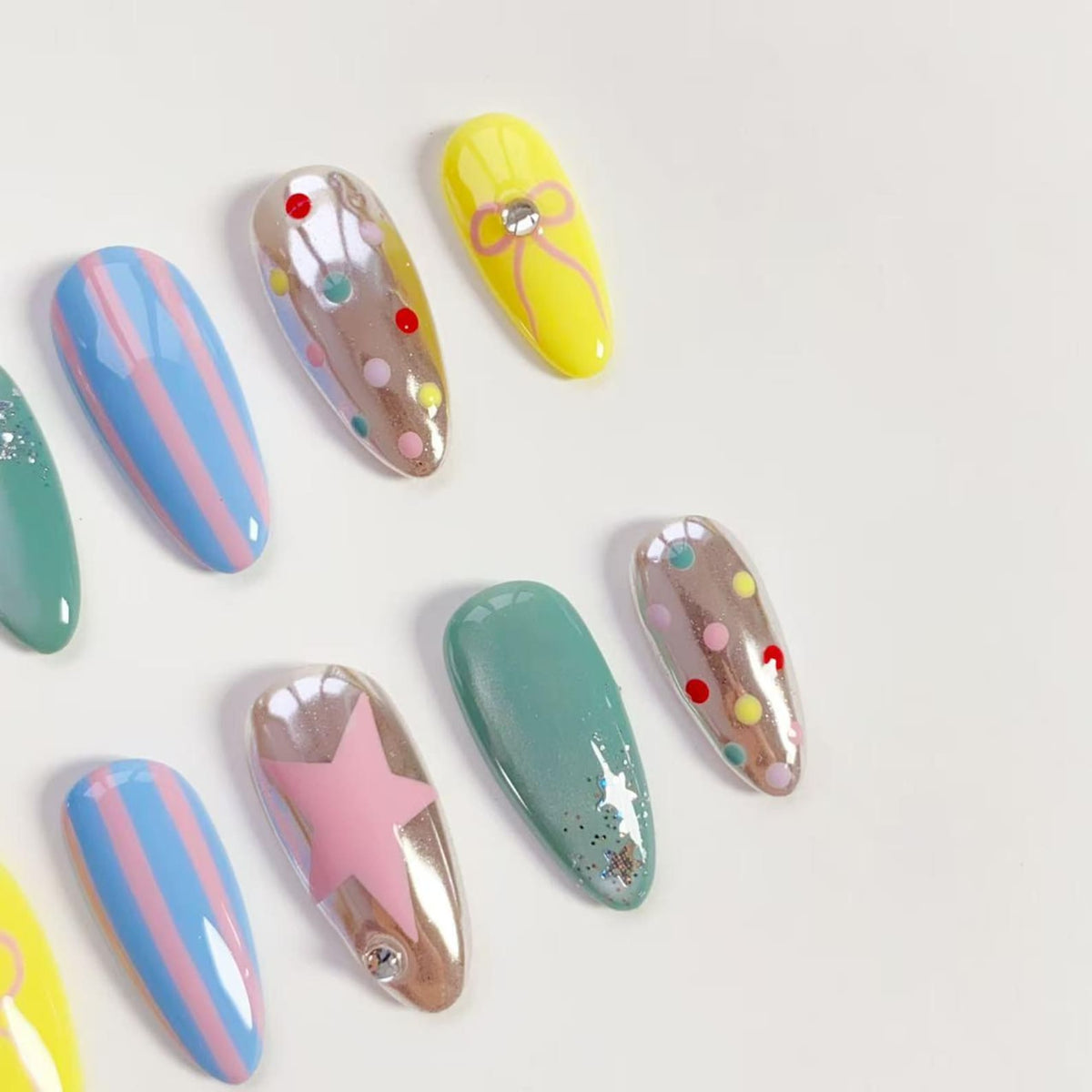 A collection of colorful, almond-shaped artificial nails. Designs include pastel blue and pink stripes, a pink star, yellow with a bow, and iridescent nails with polka dots. Some nails have glitter accents. The nails are arranged on a white surface.