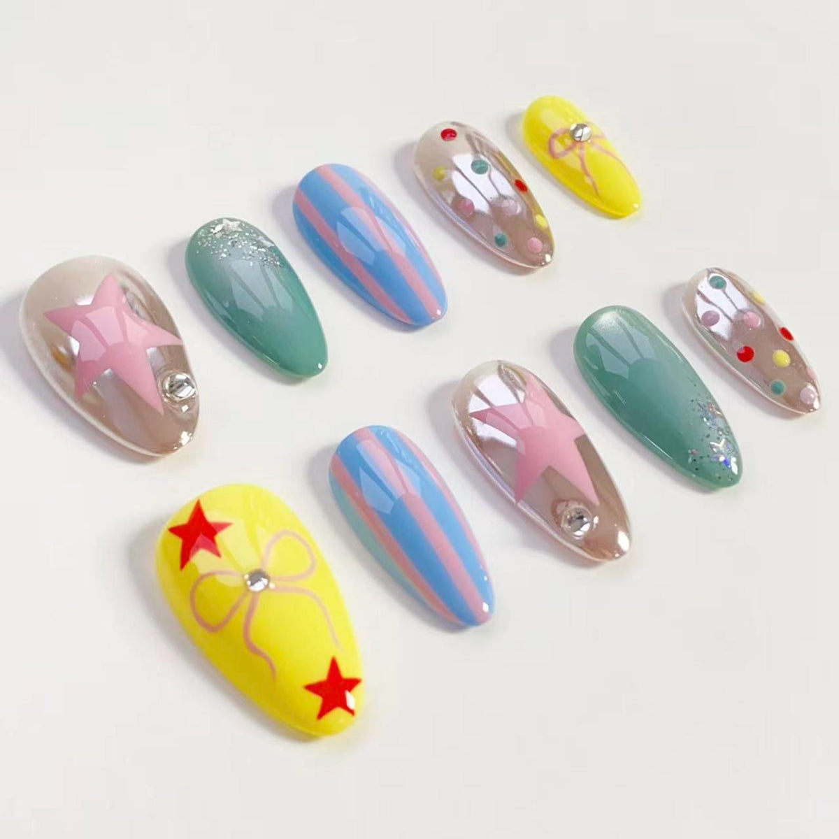 A collection of artificial nails in various designs. Some nails are yellow with red stars, others are iridescent with pink stars, and some are teal or blue with stripes. Each nail has a glossy finish.