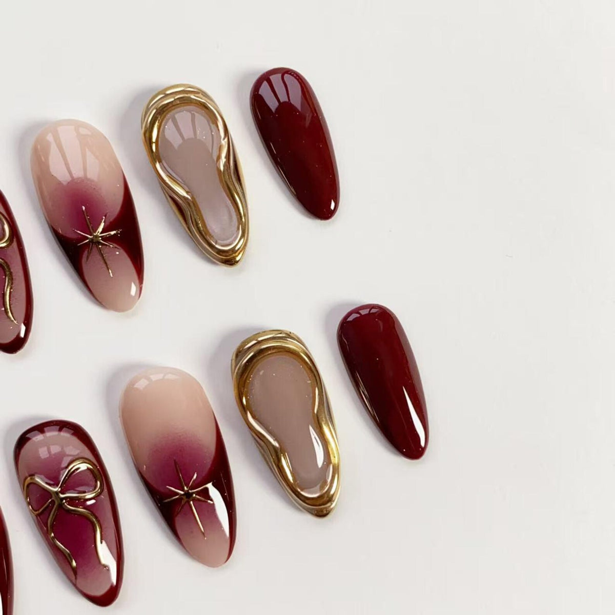 An assortment of artificial nails in various designs. Some nails feature a gradient of nude to burgundy, adorned with gold star or bow accents. Others have a solid burgundy color or a gold-framed design.