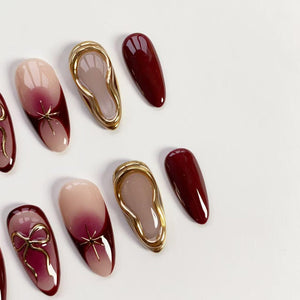 An assortment of artificial nails in various designs. Some nails feature a gradient of nude to burgundy, adorned with gold star or bow accents. Others have a solid burgundy color or a gold-framed design.
