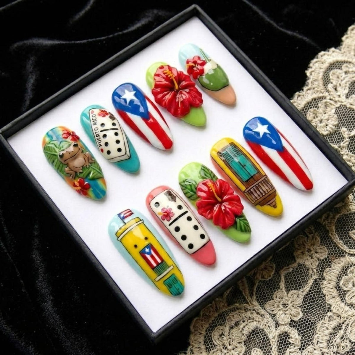 A set of ten artificial nails with various designs. The nails feature images of the Puerto Rican flag, dominoes, flowers, and a frog. The nails are displayed in a black box on a white surface.