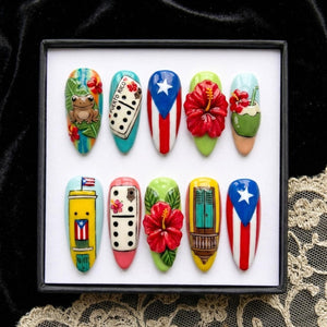 A set of ten artificial nails with various designs. The designs include a frog, dominoes, the Puerto Rican flag, a red hibiscus flower, and a coconut drink. The nails are displayed in a black box.
