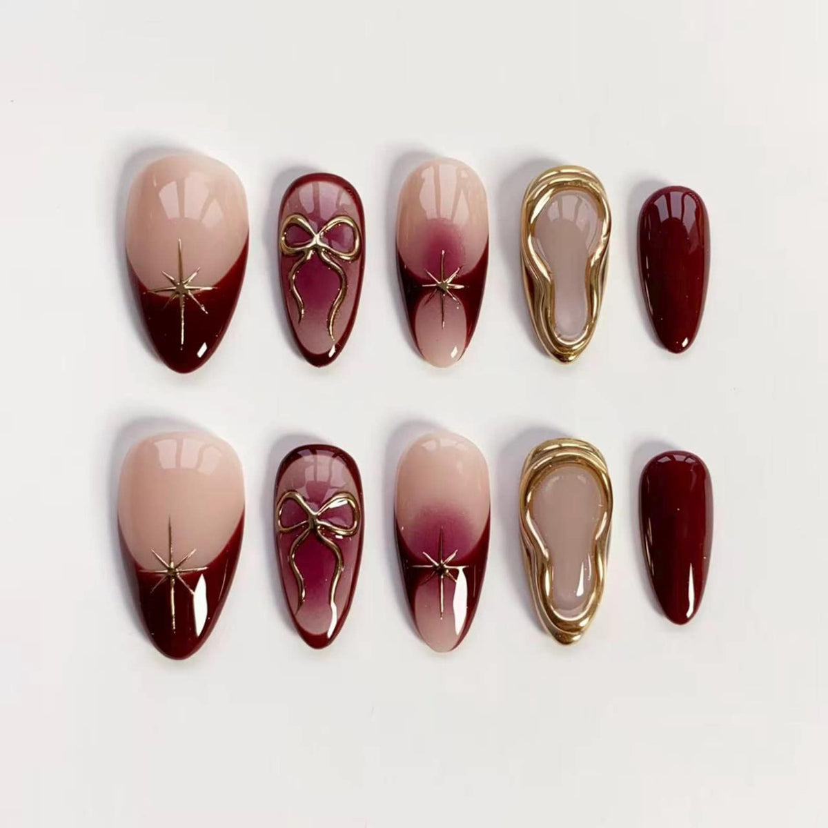 A set of ten artificial nails in various designs. The nails feature a nude base with burgundy tips, gold starburst accents, gold bow details, and gold-framed designs. One nail is solid burgundy.