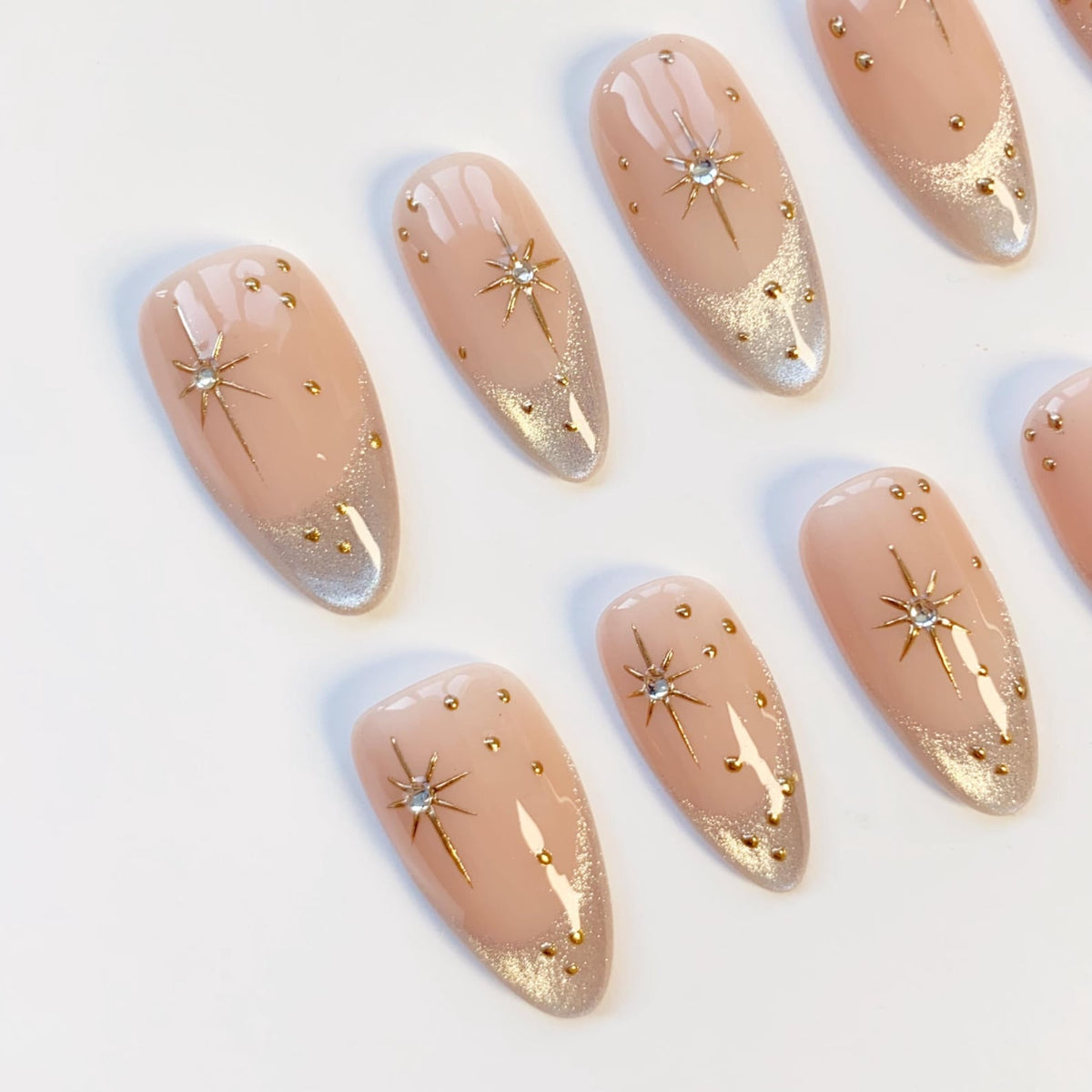 Oval-shaped artificial nails with a nude base, silver glitter tips, and gold star and dot accents. The nails are arranged on a white surface, showcasing a celestial design. The nail art features a glamorous and elegant aesthetic.