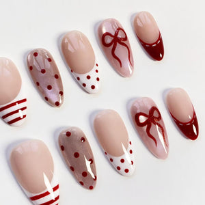 A collection of artificial nails with various designs. Some nails feature a nude base with red polka dots or a red bow. Others have a white base with red stripes or a red French tip. The nails are oval-shaped.