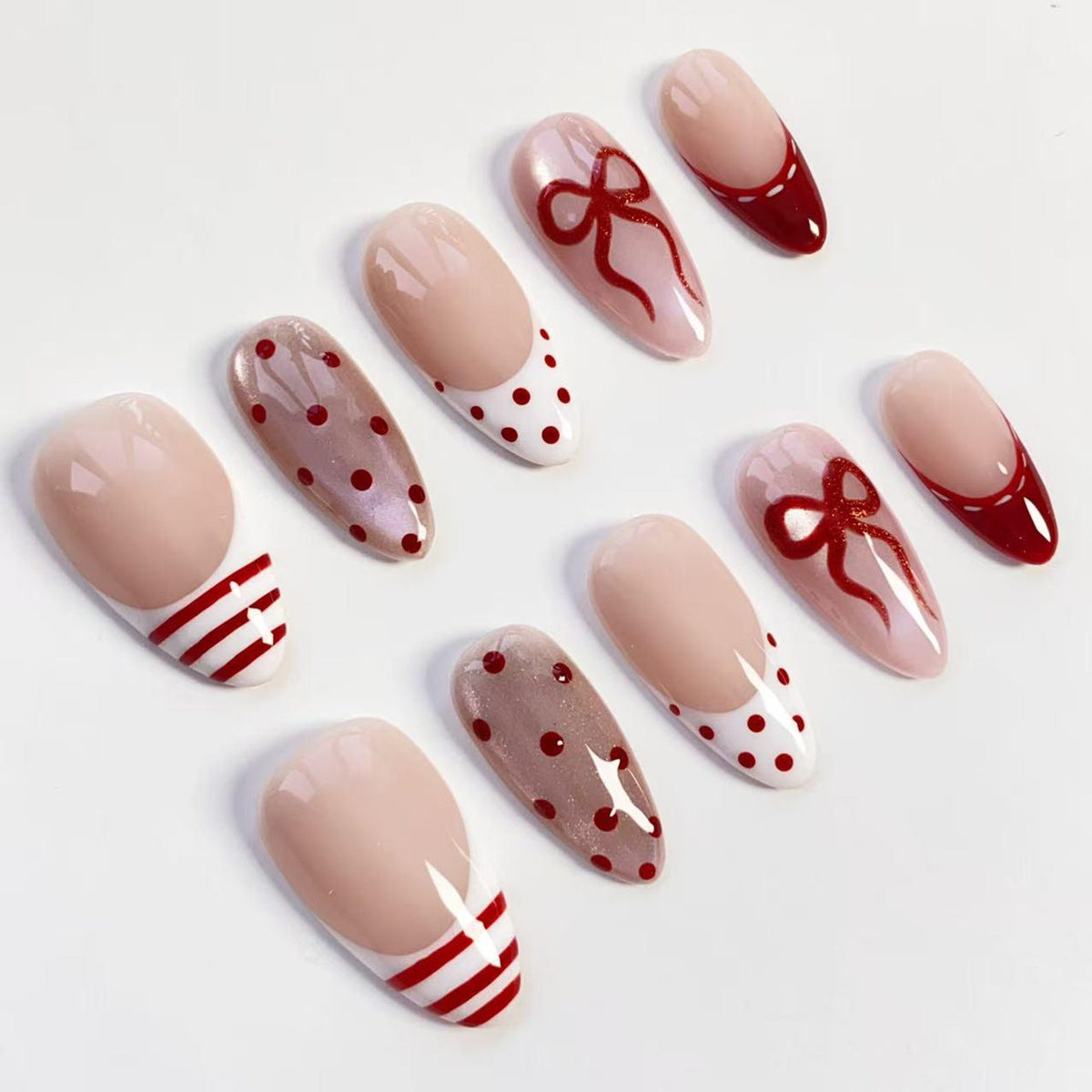 A collection of artificial nails with various designs. Some nails feature a nude base with red accents, including stripes, polka dots, and a bow. Others have a solid red tip or a nude base with a red French tip.