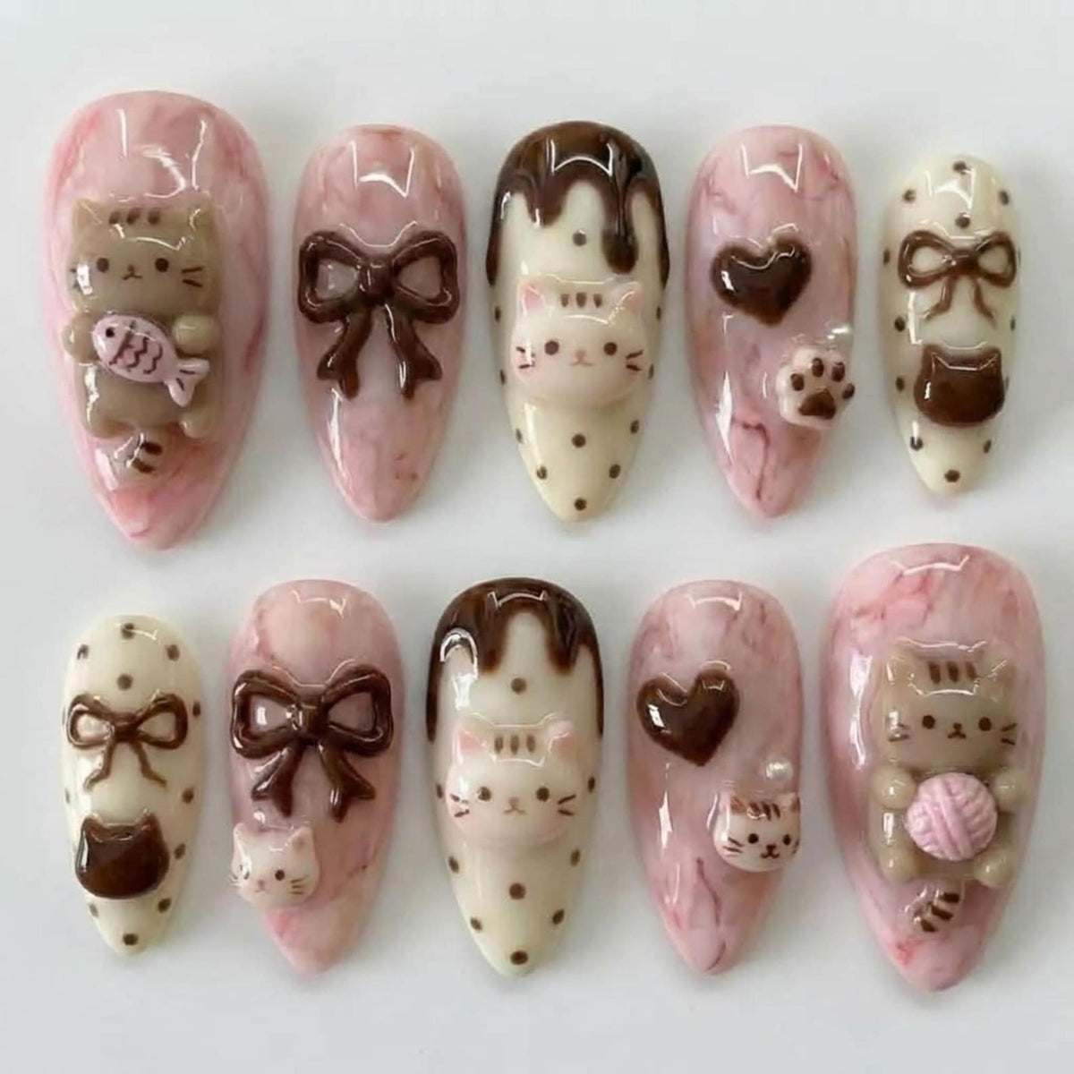 A set of ten almond-shaped false nails with a pink marble effect. Each nail features 3D embellishments, including brown bows, cat faces, hearts, paw prints, and a cat holding a ball of yarn or a fish.