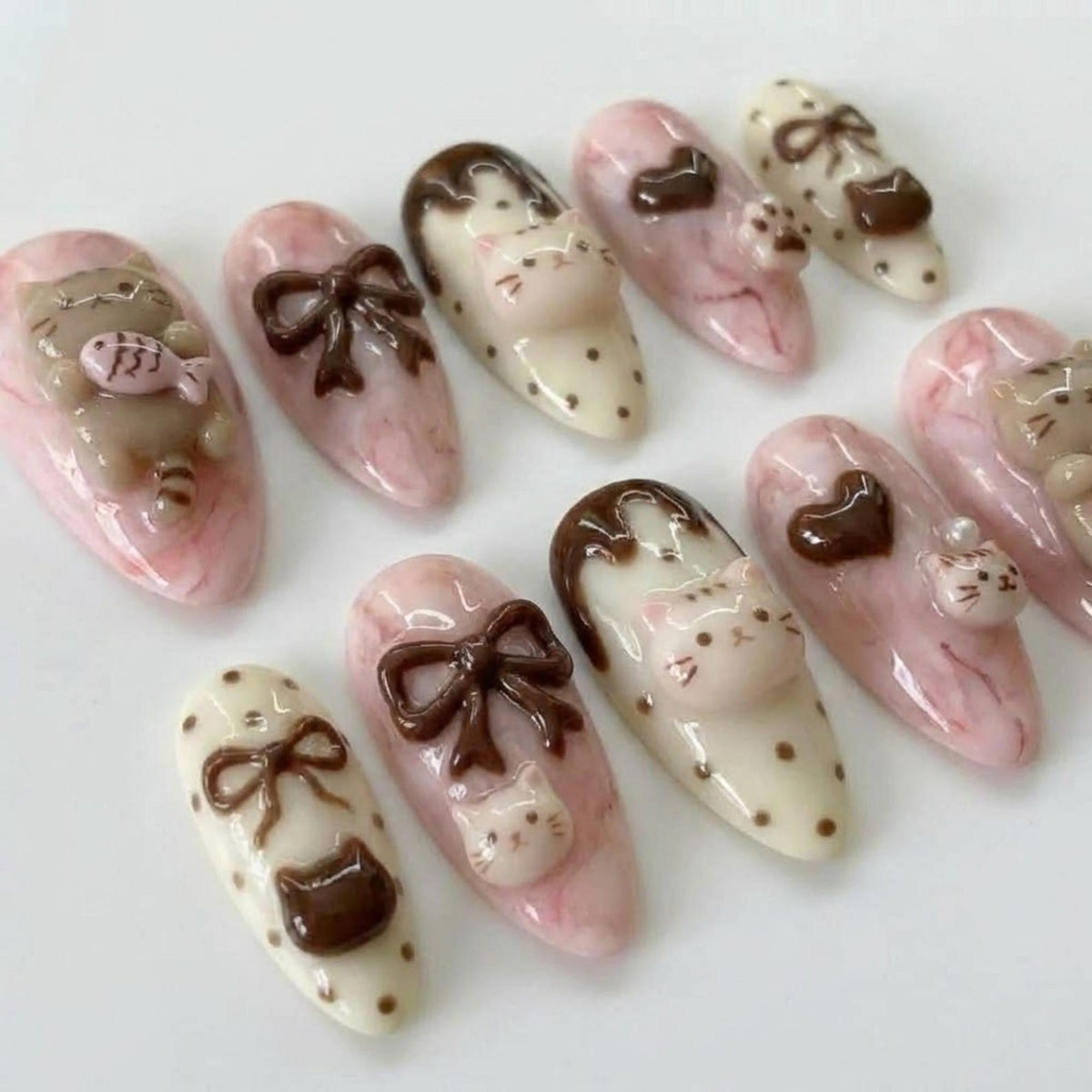 A collection of ten artificial nails with various designs. The nails feature a pink and white marbled base, adorned with 3D cat and bow designs in brown and white. Some nails have a polka dot pattern.