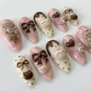 A collection of ten artificial nails with various designs. The nails feature a pink and white marbled base, adorned with 3D cat and bow designs in brown and white. Some nails have a polka dot pattern.