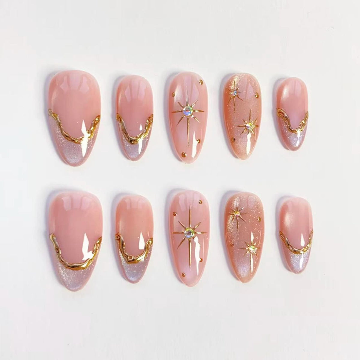 Set of ten almond-shaped false nails. The nails are a light pink with gold accents. Some nails feature a gold starburst design with a central rhinestone. Others have a gold curved design at the base.