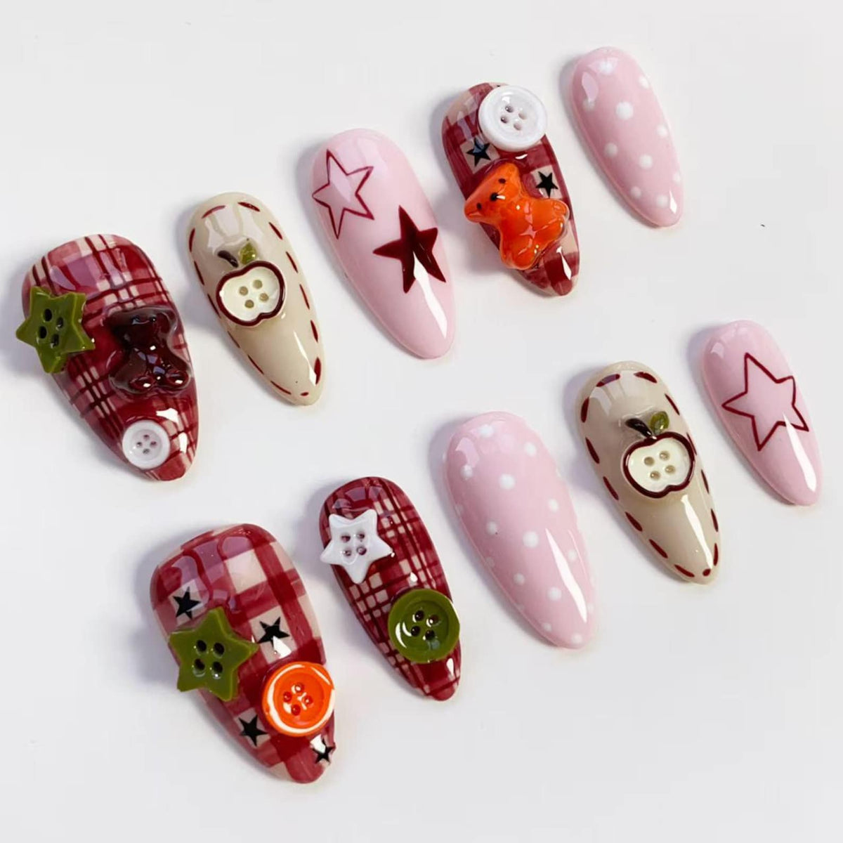 A collection of almond-shaped false nails with various designs. Some nails feature a red plaid pattern, while others are solid pink with white polka dots. Embellishments include star and apple designs, buttons, and a small orange teddy bear.
