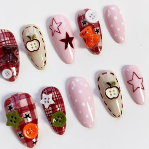 A collection of ten almond-shaped artificial nails with various designs. Some nails feature pink polish with white polka dots or star outlines. Others have plaid patterns, apple and teddy bear embellishments, and decorative buttons in green, white, a