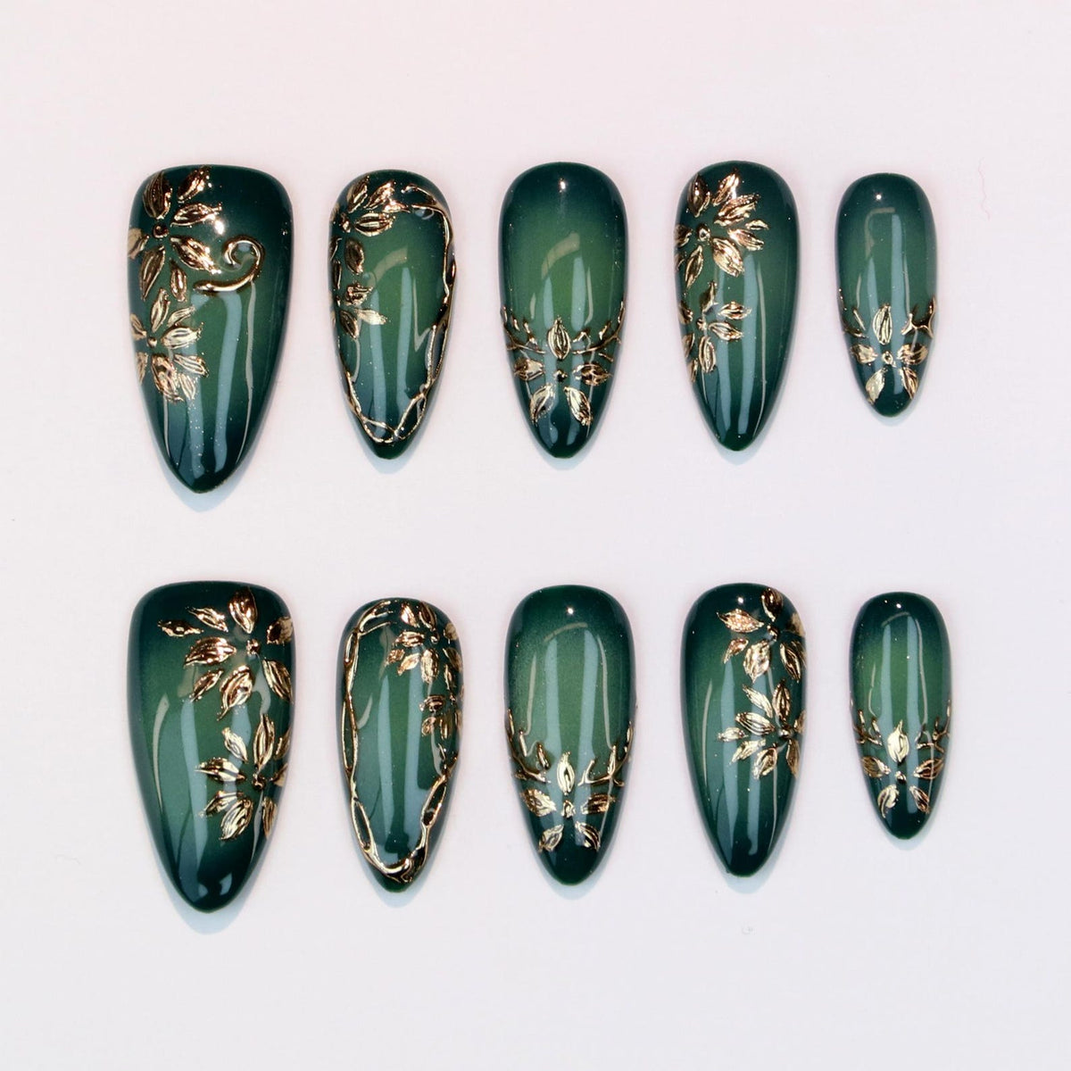 Set of ten almond-shaped false nails in a deep green hue. Each nail features gold floral designs and accents. The nails are arranged on a white surface, showcasing the intricate details of the nail art.