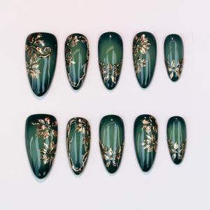 Set of ten almond-shaped false nails in a deep green hue. Each nail features gold floral designs and accents. The nails are arranged on a white surface, showcasing the intricate details of the nail art.