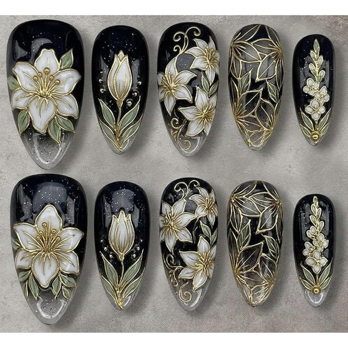 A set of ten artificial nails with a black base, featuring intricate floral designs. The designs include white flowers with gold accents, green leaves, and a clear, glittery base.