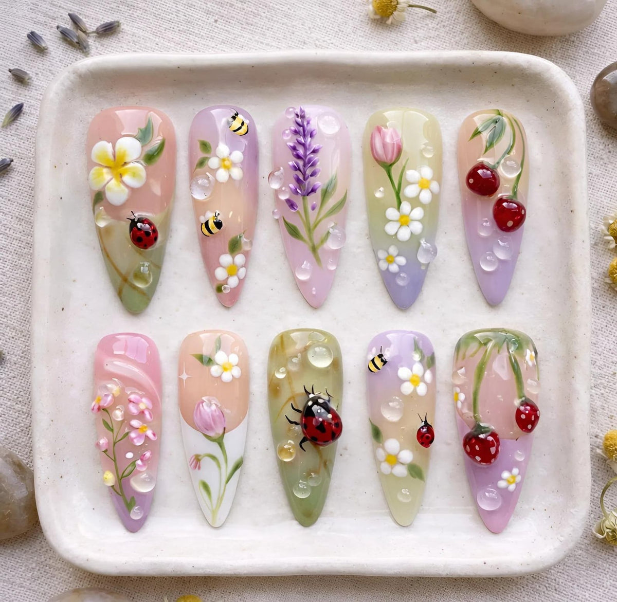 A set of ten almond-shaped, painted artificial nails. Each nail features a unique design, including flowers, bees, ladybugs, and cherries, with water droplet accents. The color palette includes pinks, greens, purples, and yellows.